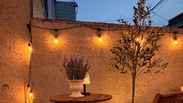A patio with two wooden chairs, a small table, a potted plant, and string lights against a textured wall. - River Place -Walk to Jefferson Methodist, 2BR Queen Beds 1 Bath -Patio & Grill (Philadelphia)