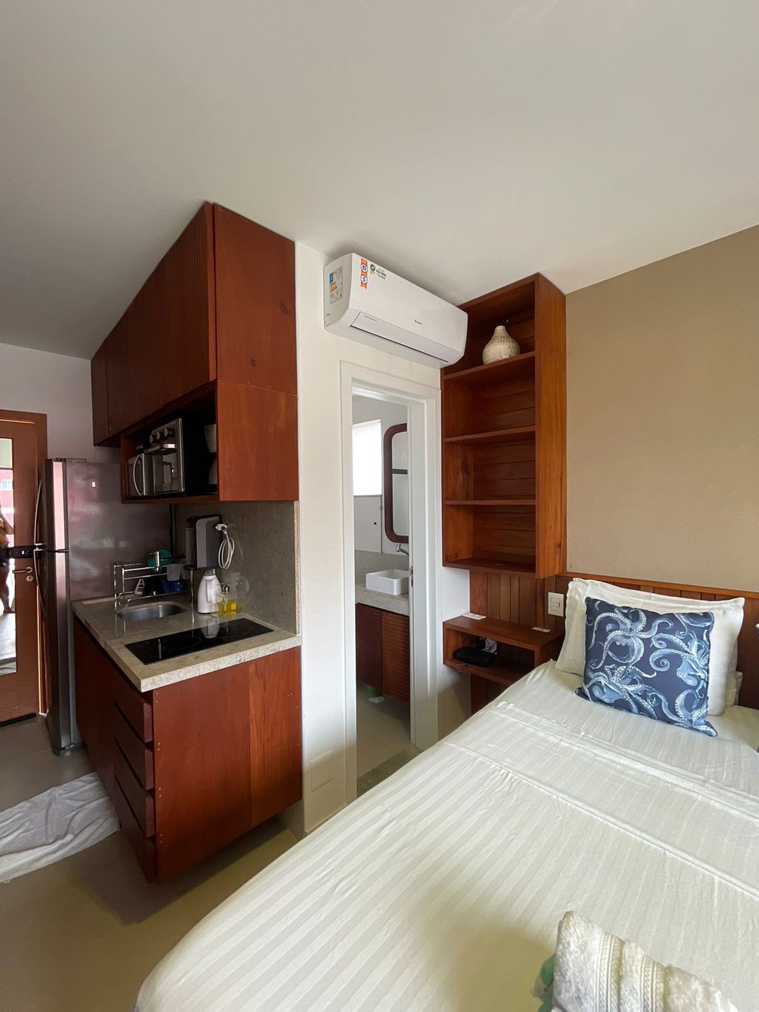Quarto de hotel compacto com cozinha compacta, cama com travesseiro estampado e pequena mesa de cabeceira.