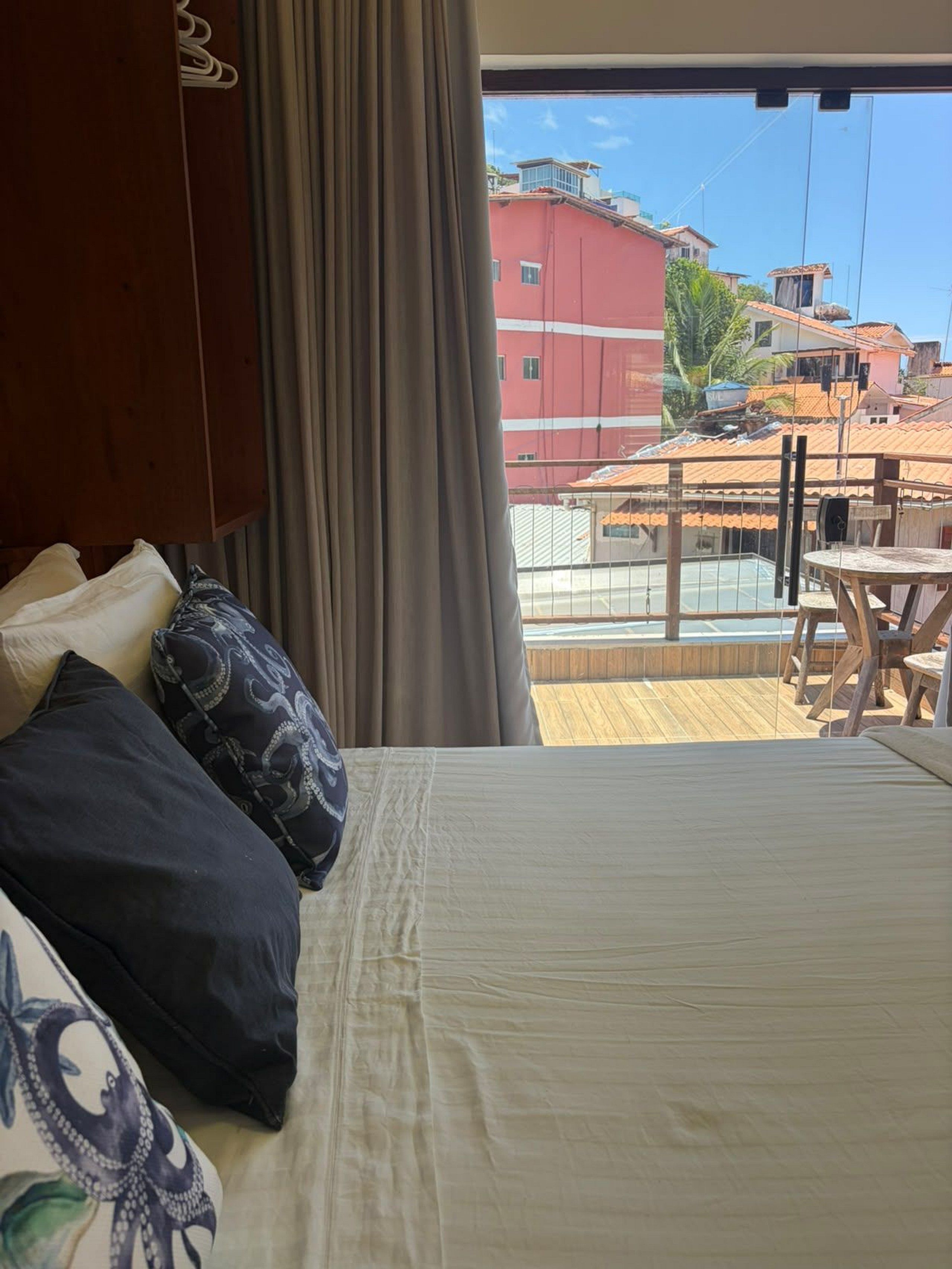 A hotel room with a bed, a wooden wardrobe, a view of the cityscape, and a balcony with a table and chairs.