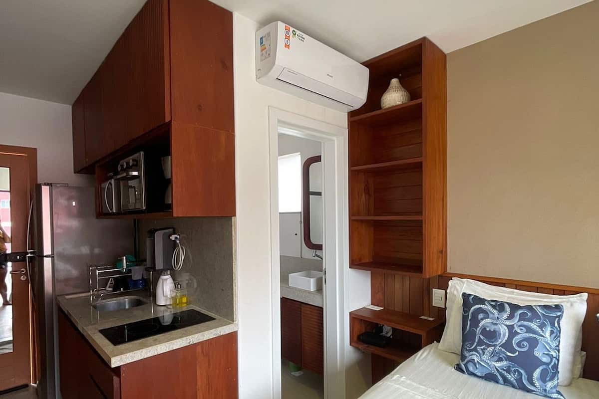 A compact hotel room with a kitchenette, a bed with a patterned pillow, and a small bedside table.