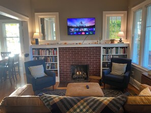Ein Wohnzimmer mit Kamin, zwei Sesseln, einer Couch und einem Fernseher, der über dem Kamin angebracht ist, sowie Bücherregalen an beiden Seiten.