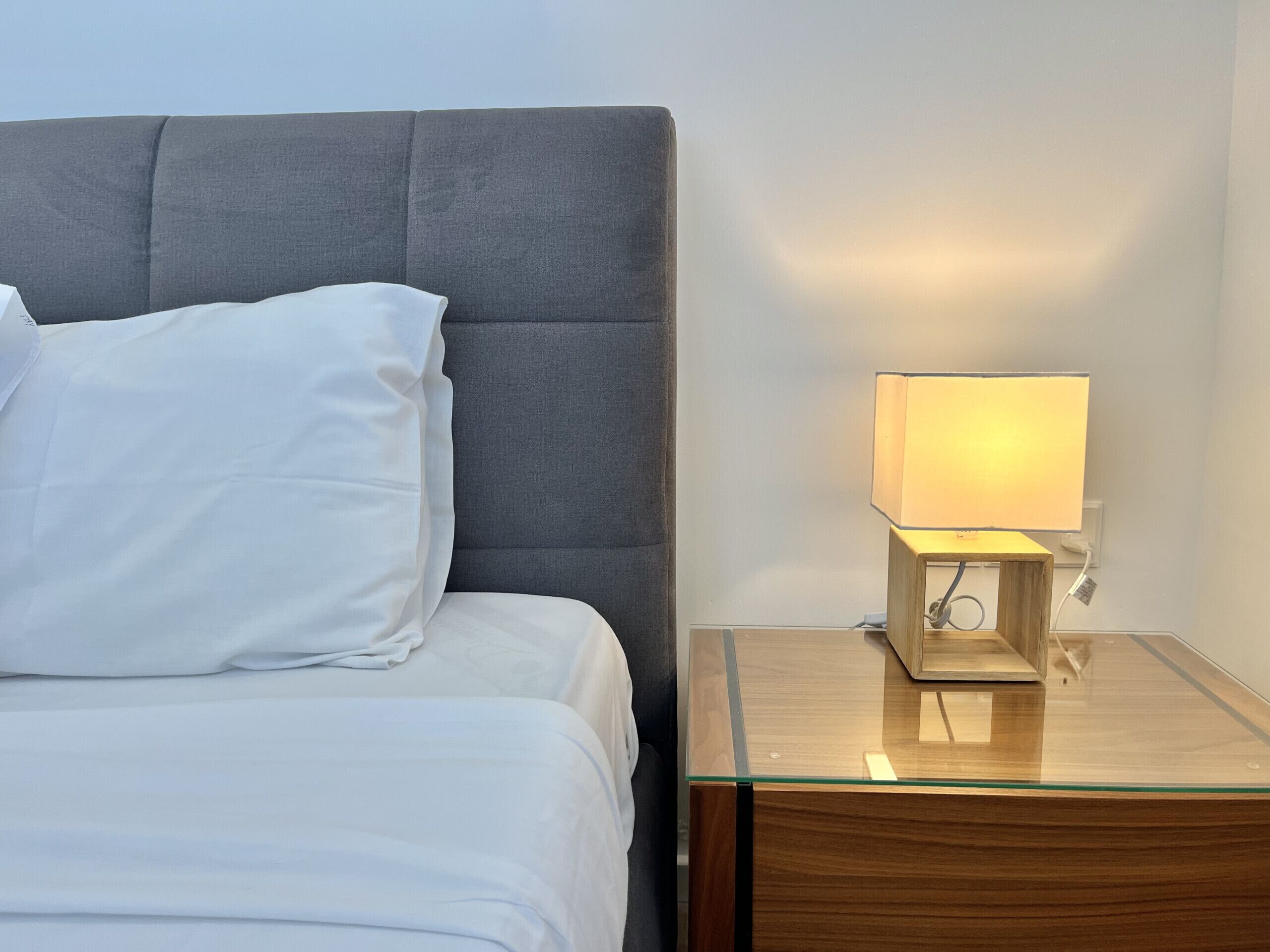 A bed with white bedding, a grey upholstered headboard, and a bedside table with a lamp.