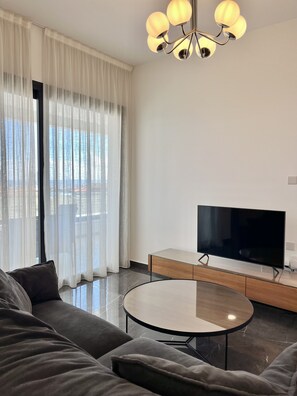 A modern living room with a sofa, a TV, a glass coffee table, and a chandelier. - STAY Anna Residence Sea View Apartment 201 (Agios Athanasios)