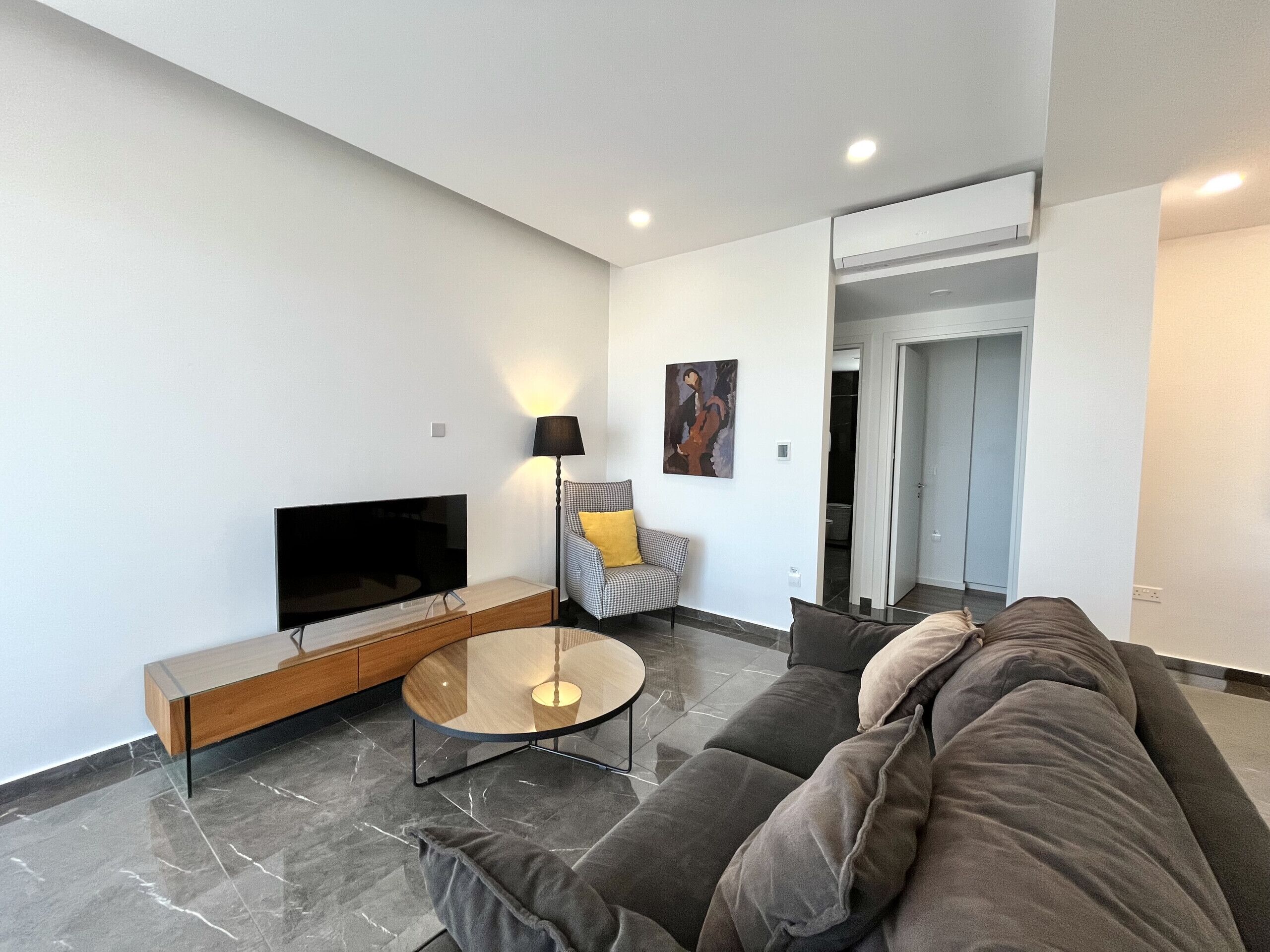 Ein modernes Wohnzimmer mit Flachbildfernseher, Glastisch, Sofa, Sessel und Lampe.