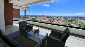 En balkon med fletmøbler, et glasbord og udsigt over by og hav.