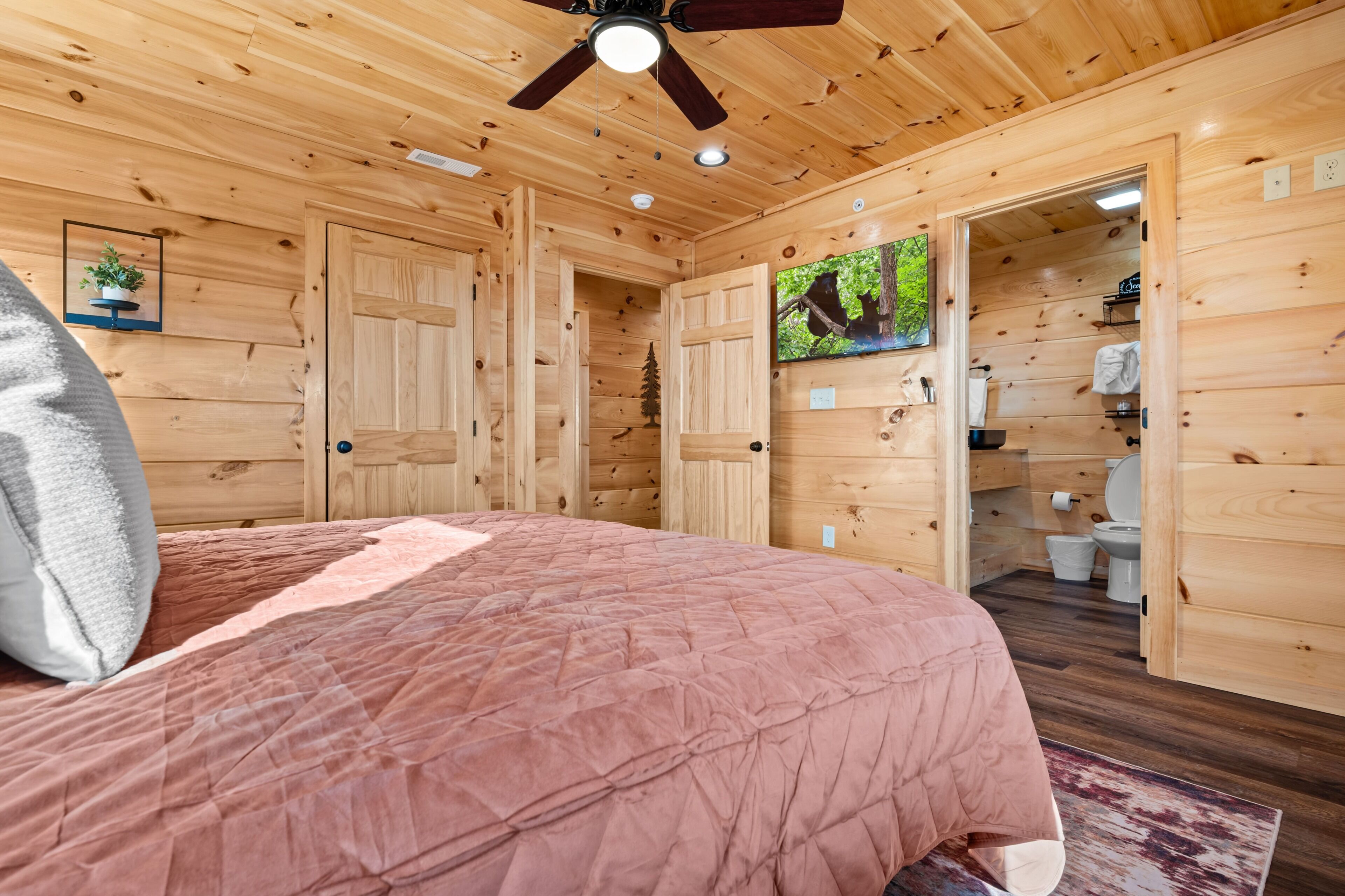 A wooden cabin bedroom with a bed, ceiling fan, and a bathroom visible through an open door.