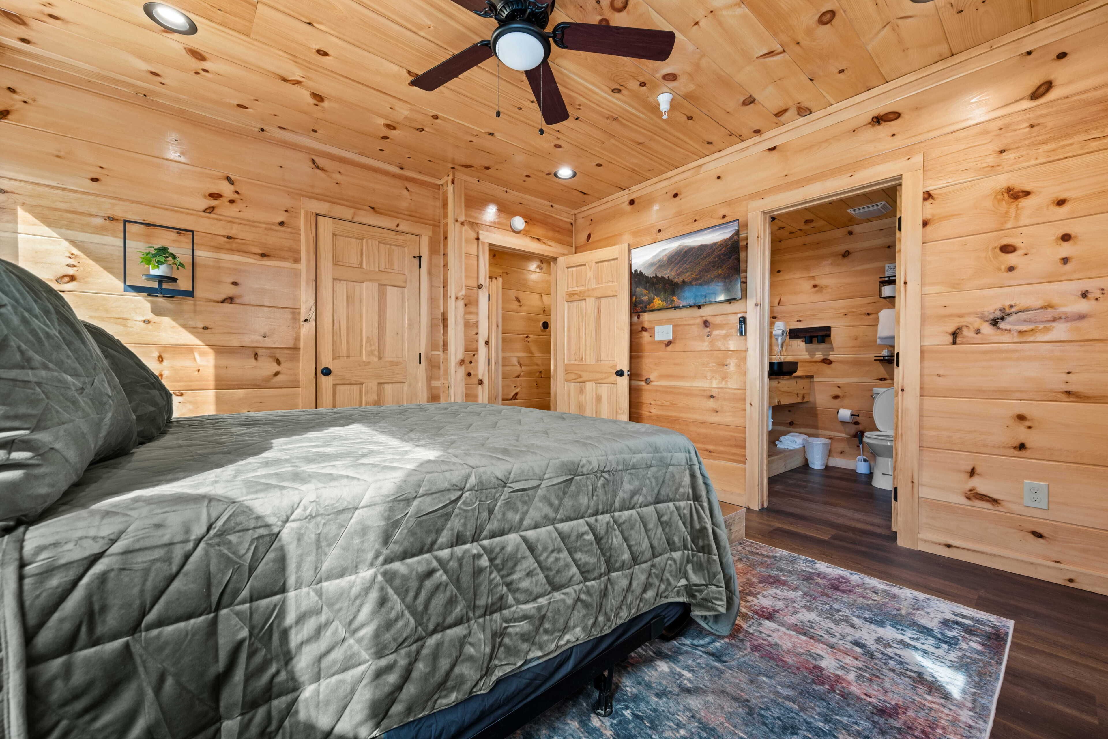 A wooden cabin interior with a bed, ceiling fan, and a bathroom visible through an open door.