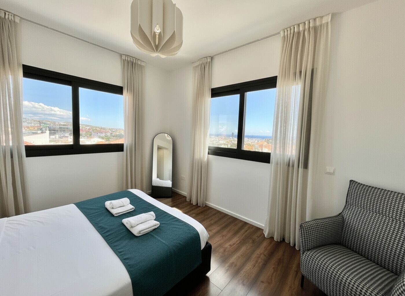 Ein Hotelzimmer mit Bett, Stuhl, Spiegel und großen Fenstern mit Blick auf die Stadt.
