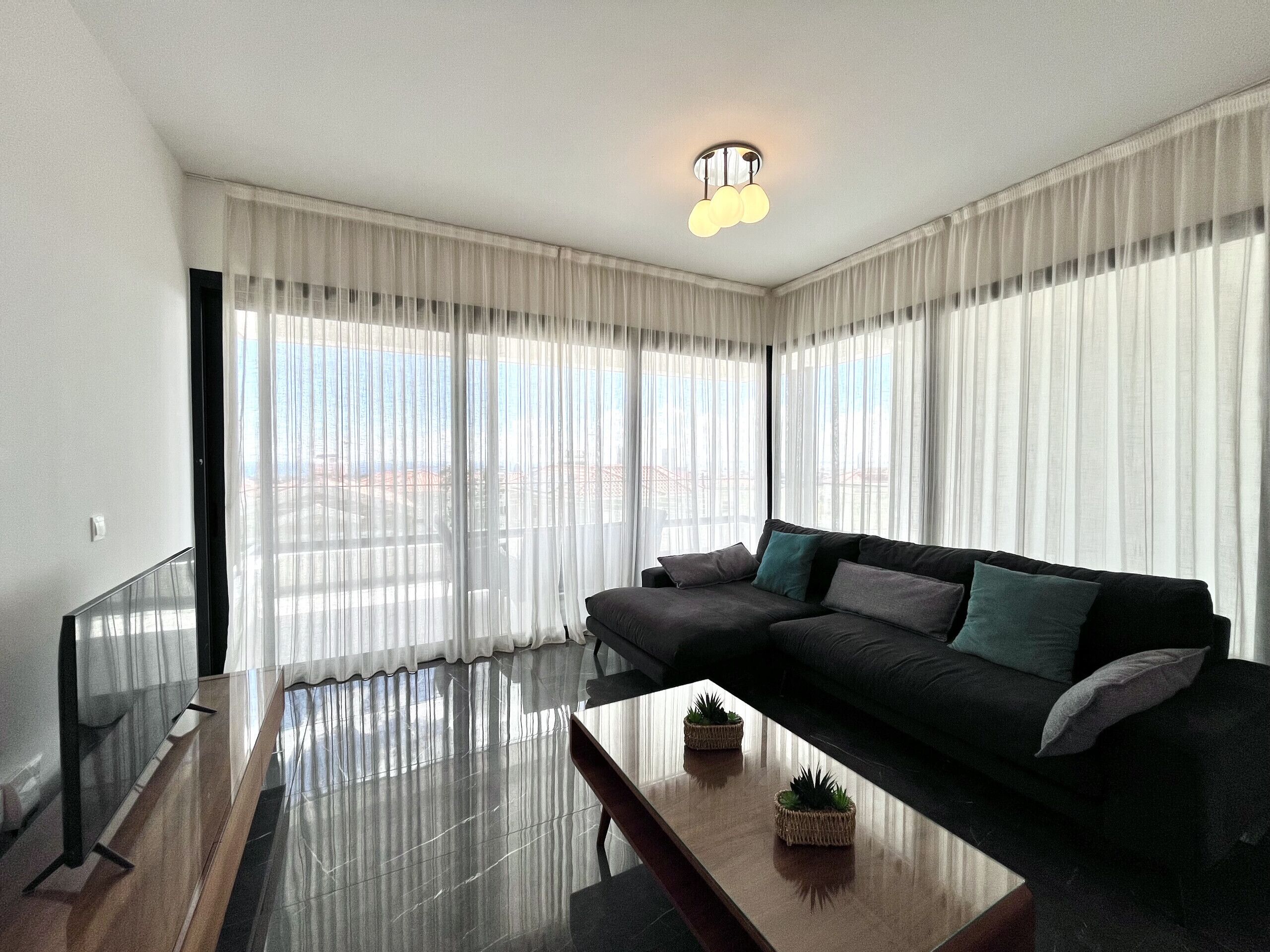 Ein modernes Wohnzimmer mit einem schwarzen Sofa, einem Glastisch und transparenten Vorhängen.