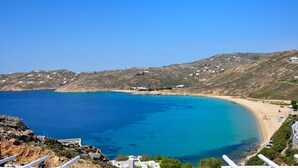 Un paisaje costero con mar azul turquesa, playa arenosa y colinas al fondo donde se ven algunas edificaciones dispersas.