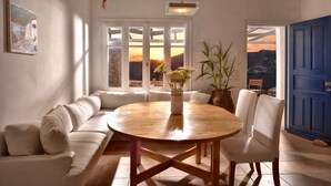 Una habitación con una mesa redonda de madera, asientos acolchados blancos, una planta en maceta y vistas a través de una puerta.