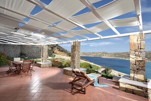 Patio esterno con pilastri in pietra, tavolo con sedie, una poltrona prendisole e vista sul mare.