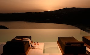 Evigheds-pool med liggestole, et bord med en drink og udsigt til solnedgang over vandet.