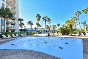 A swimming pool with water jets, surrounded by palm trees and lounge chairs, with a building in the background. - The Palms of Destin 1111 by Compass Resorts (Destin)