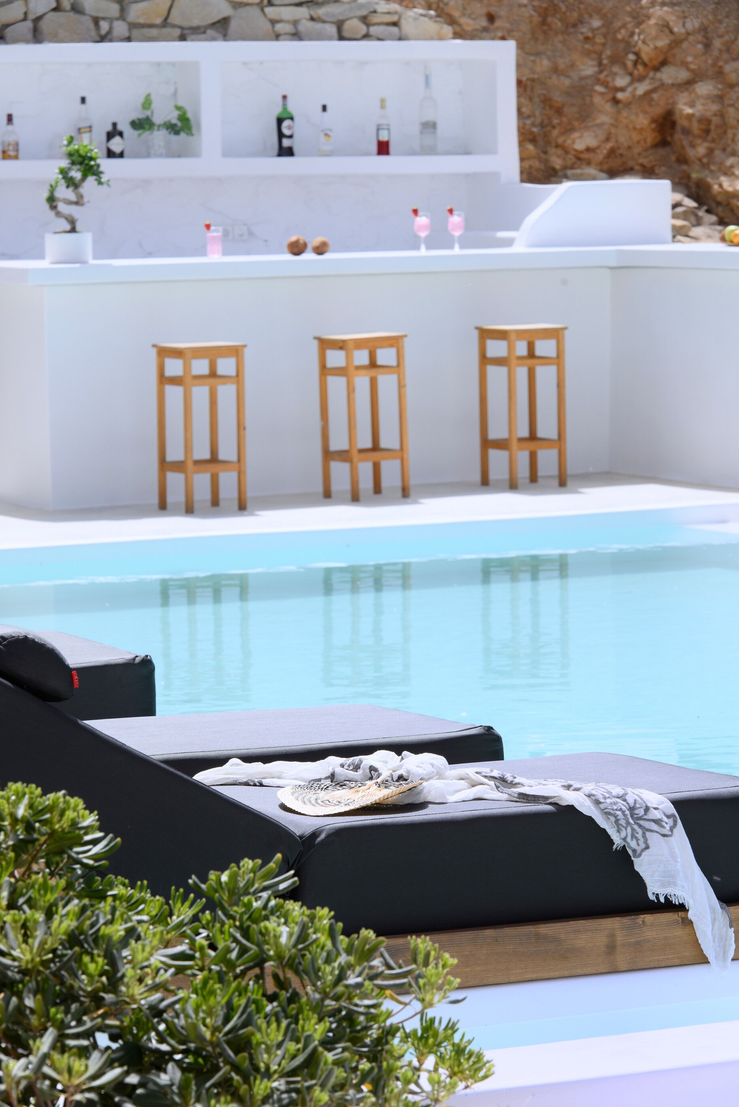 Zona de piscina con sillones reclinables, un bar con taburetes y un muro de piedra de fondo.