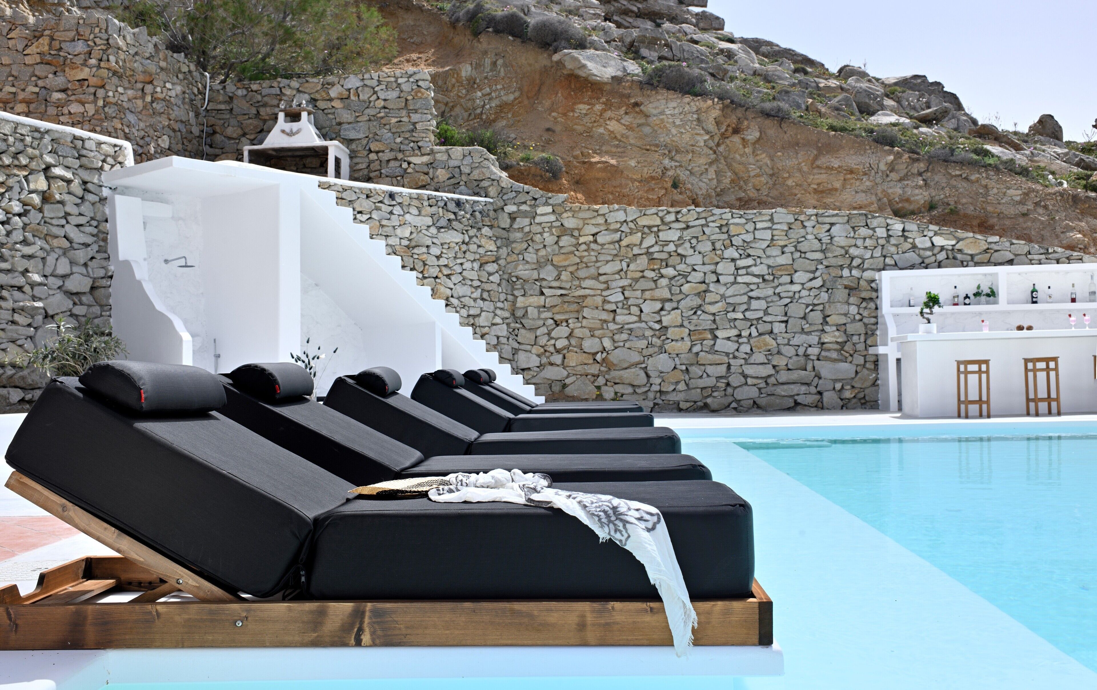 Un espace de détente aménagé au bord de la piscine, avec des chaises longues noires, un mur en pierre et un bâtiment blanc en arrière-plan.