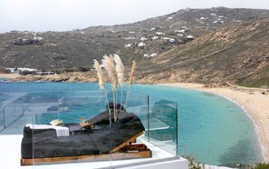 Une piscine à débordement entièrement vitrée, offrant une vue sur un paysage côtier, notamment une plage et des bâtiments blancs situés sur une colline.