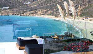 Une piscine à débordement en verre offrant une vue sur une plage et des montagnes, un canapé noir et des herbes des pampas.