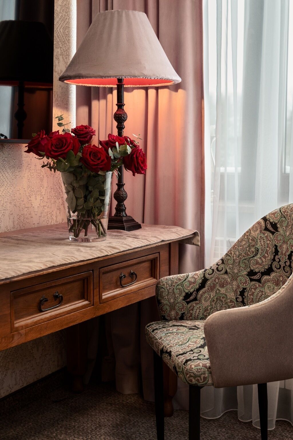 A wooden desk with a vase of red roses, a lamp, and a patterned chair.