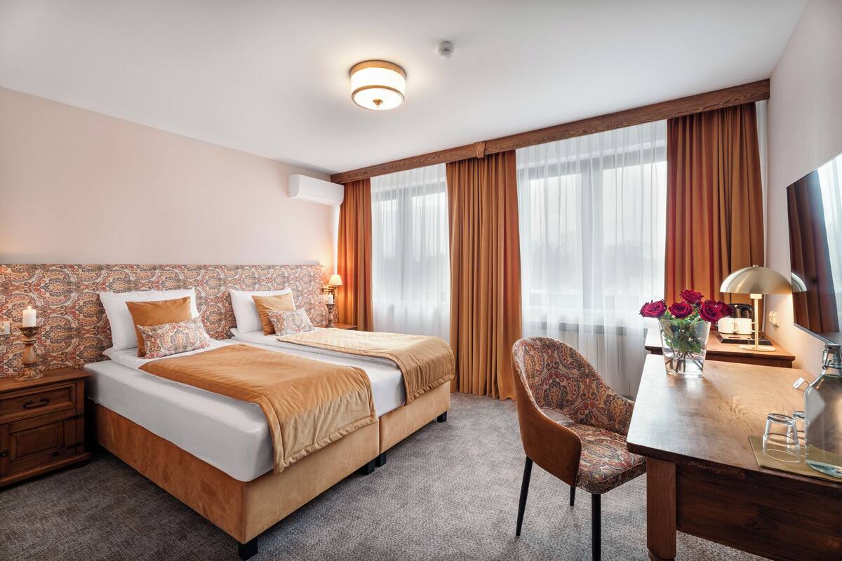 A hotel room with a double bed, a desk with a chair, a TV, and a vase with flowers.