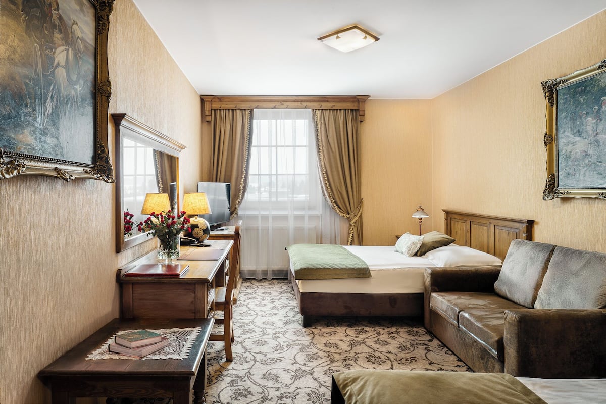 A hotel room with a bed, a sofa, a desk with a lamp, a mirror, and a painting on the wall.
