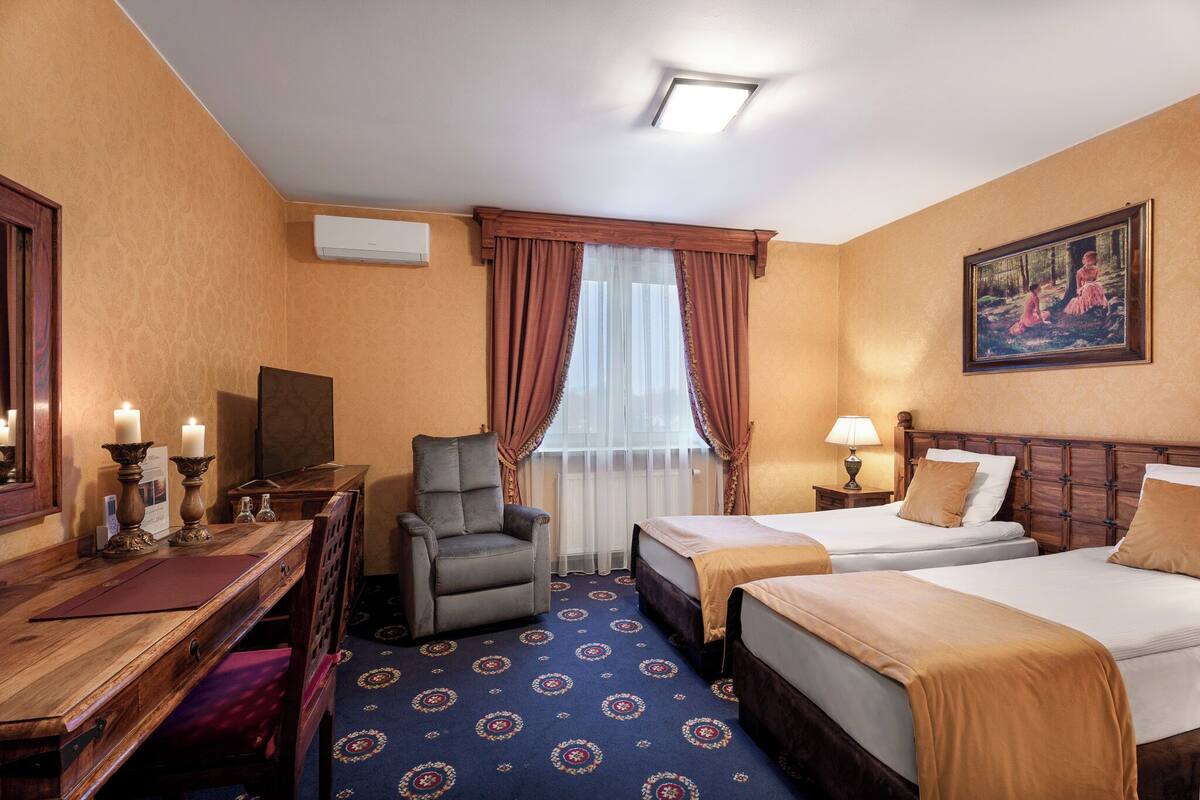 A hotel room with two beds, a desk, a chair, a window with curtains, a painting on the wall, and an air conditioning unit.