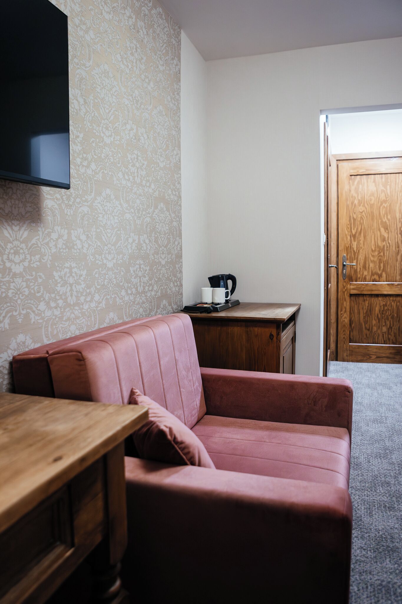 A room with a wooden desk, a pink sofa, a wooden door, and a wall-mounted TV.