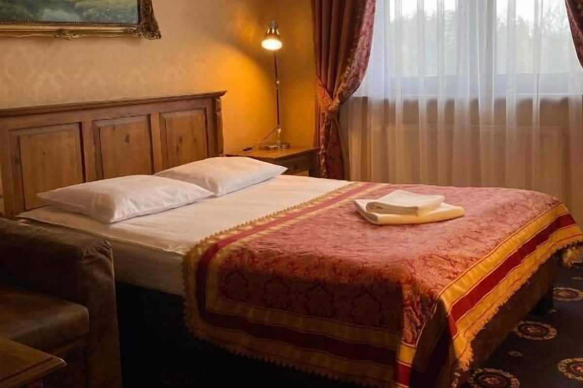 A hotel room with a wooden bed, a nightstand with a lamp, a painting on the wall, and a window with curtains.