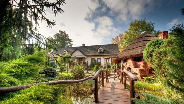 Een houten bruggetje naar een landelijk huis met rieten dak, omgeven door weelderige begroeiing en een vijver.