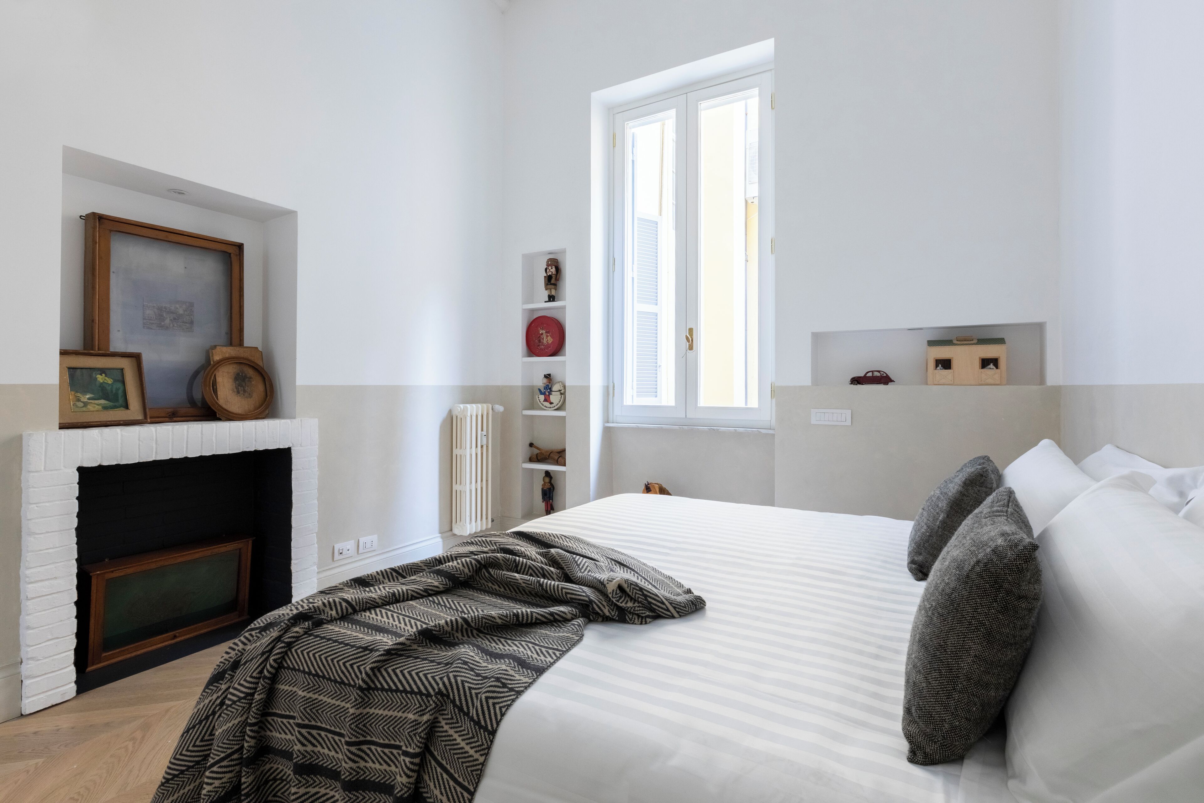 A bedroom with a white brick fireplace, a large bed with grey pillows, a window with white shutters, and a shelf with decorative items.