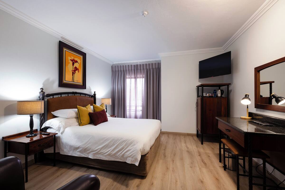 A hotel room with a bed, bedside table, lamp, desk, chair, TV, and a large mirror.