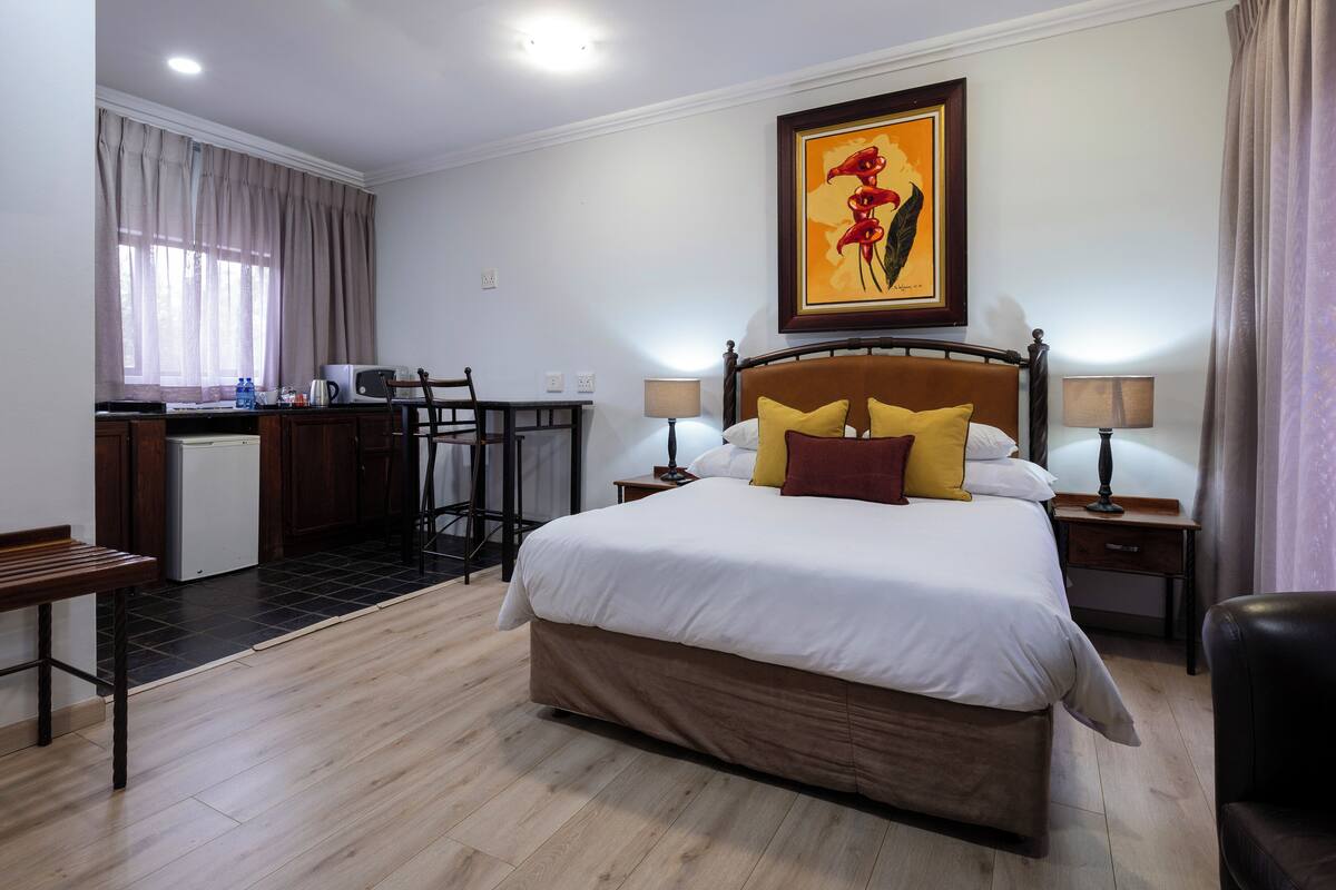 A hotel room with a bed, bedside tables, a desk with a chair, a small kitchenette, and a window with curtains.
