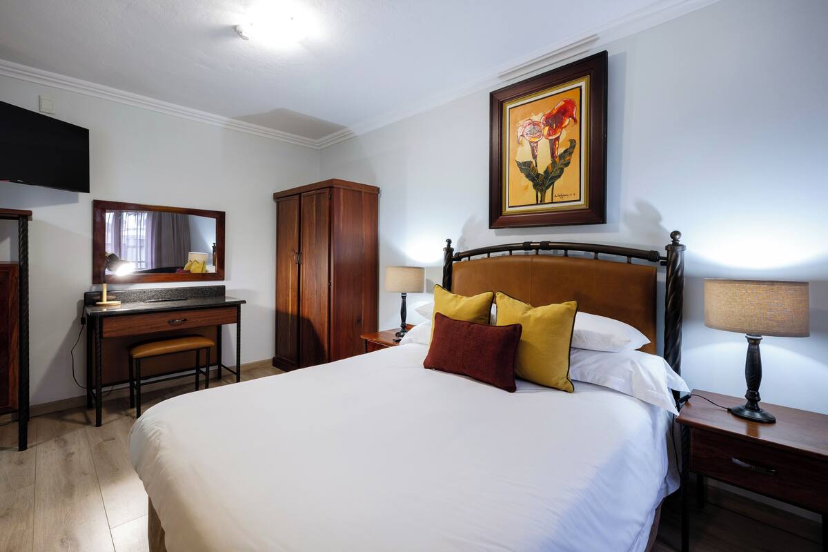 A hotel room with a large bed, a desk with a chair, a wardrobe, a bedside table with a lamp, and a painting on the wall.