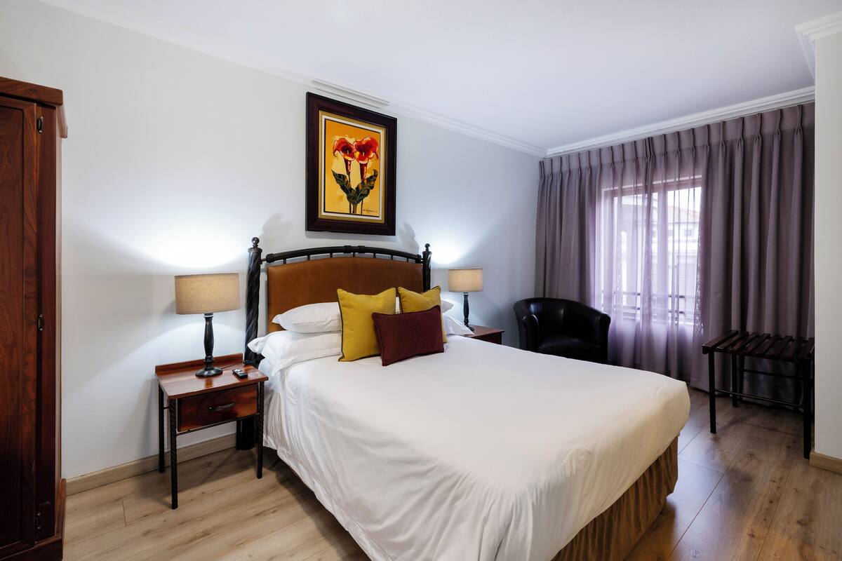 A hotel room with a large bed, two bedside tables, a chair, a window with curtains, and a painting on the wall.