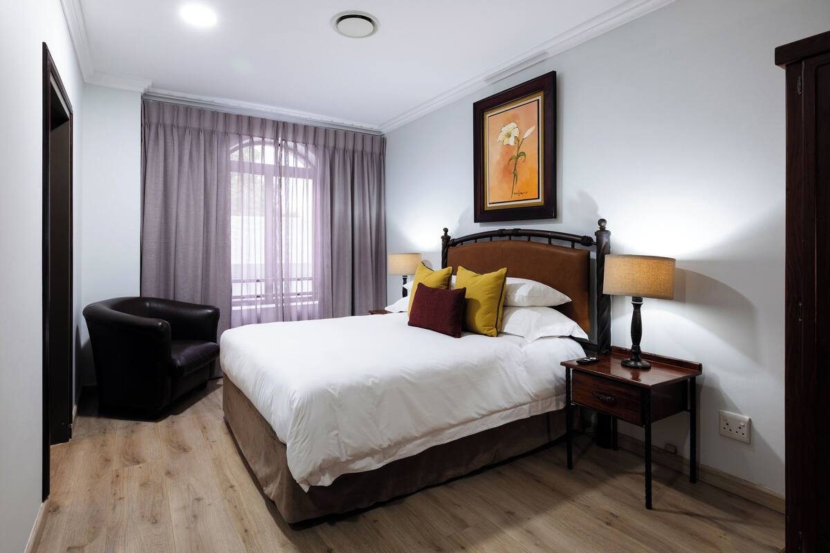 A bedroom with a bed, a chair, a nightstand, a lamp, a painting, and a window with curtains.