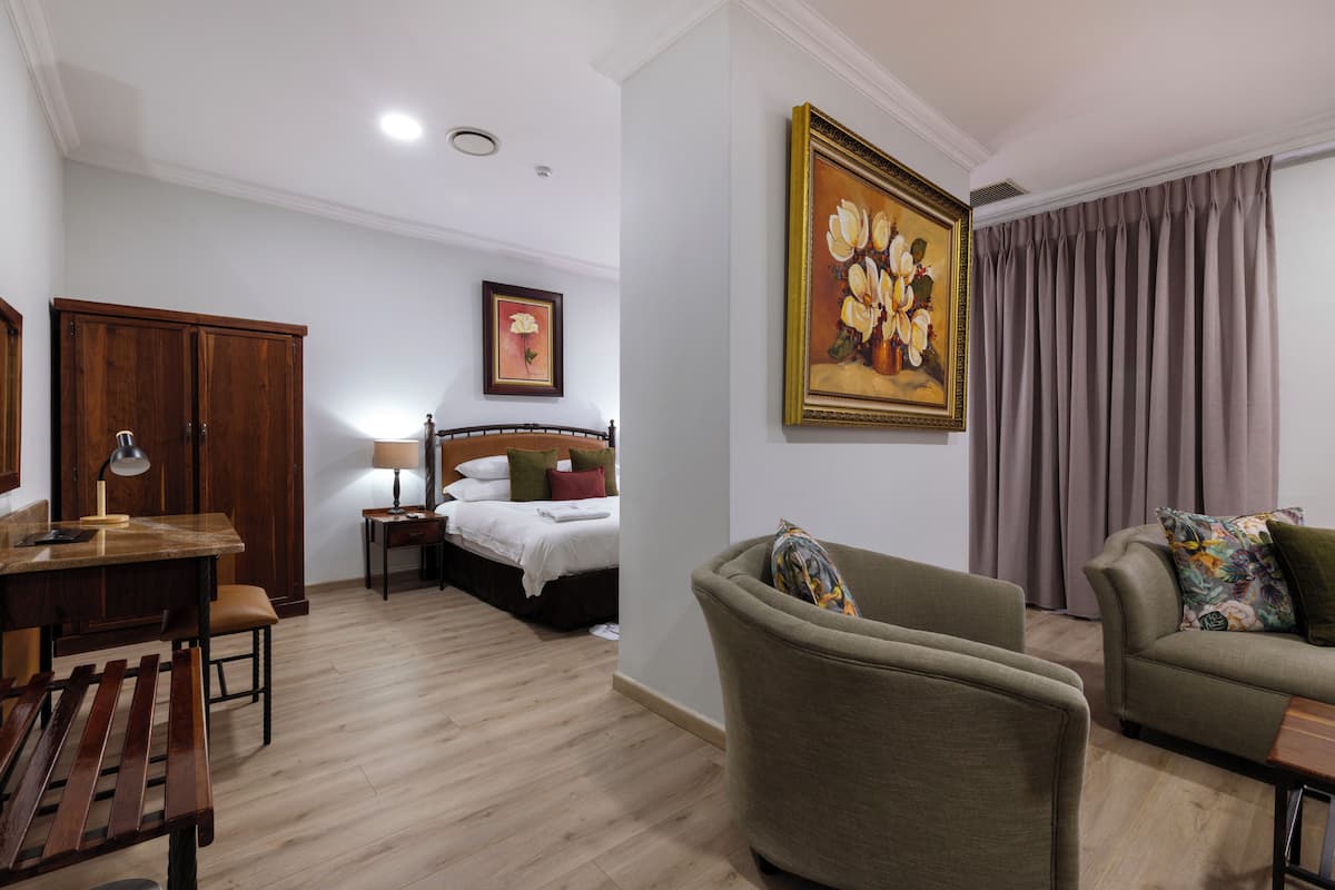 A hotel room with a wooden floor, a bed with white linens, a desk with a lamp, a chair, and a sofa with a floral pillow.
