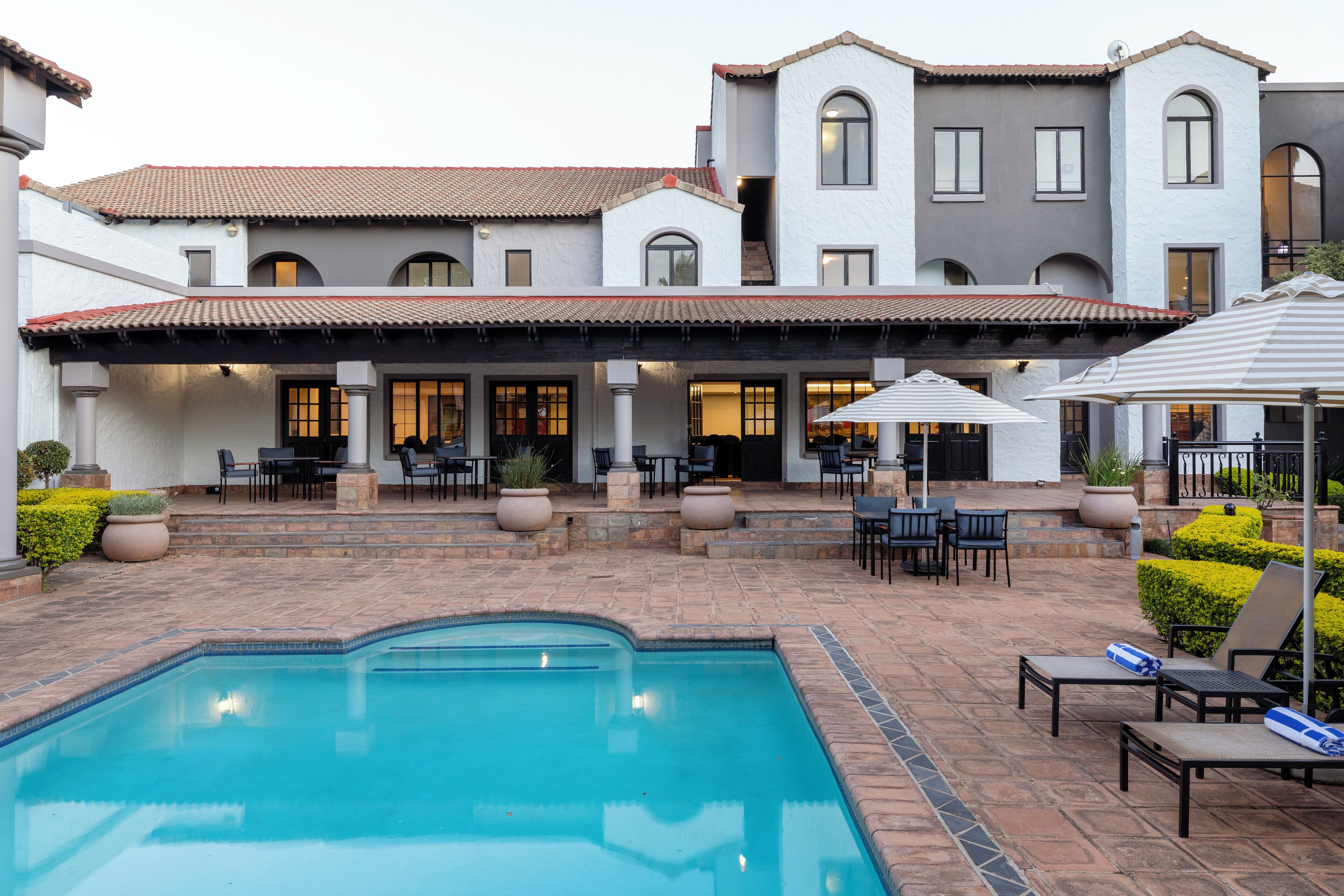 A pool area with a tiled deck, outdoor seating, and a building with multiple windows and a covered patio.