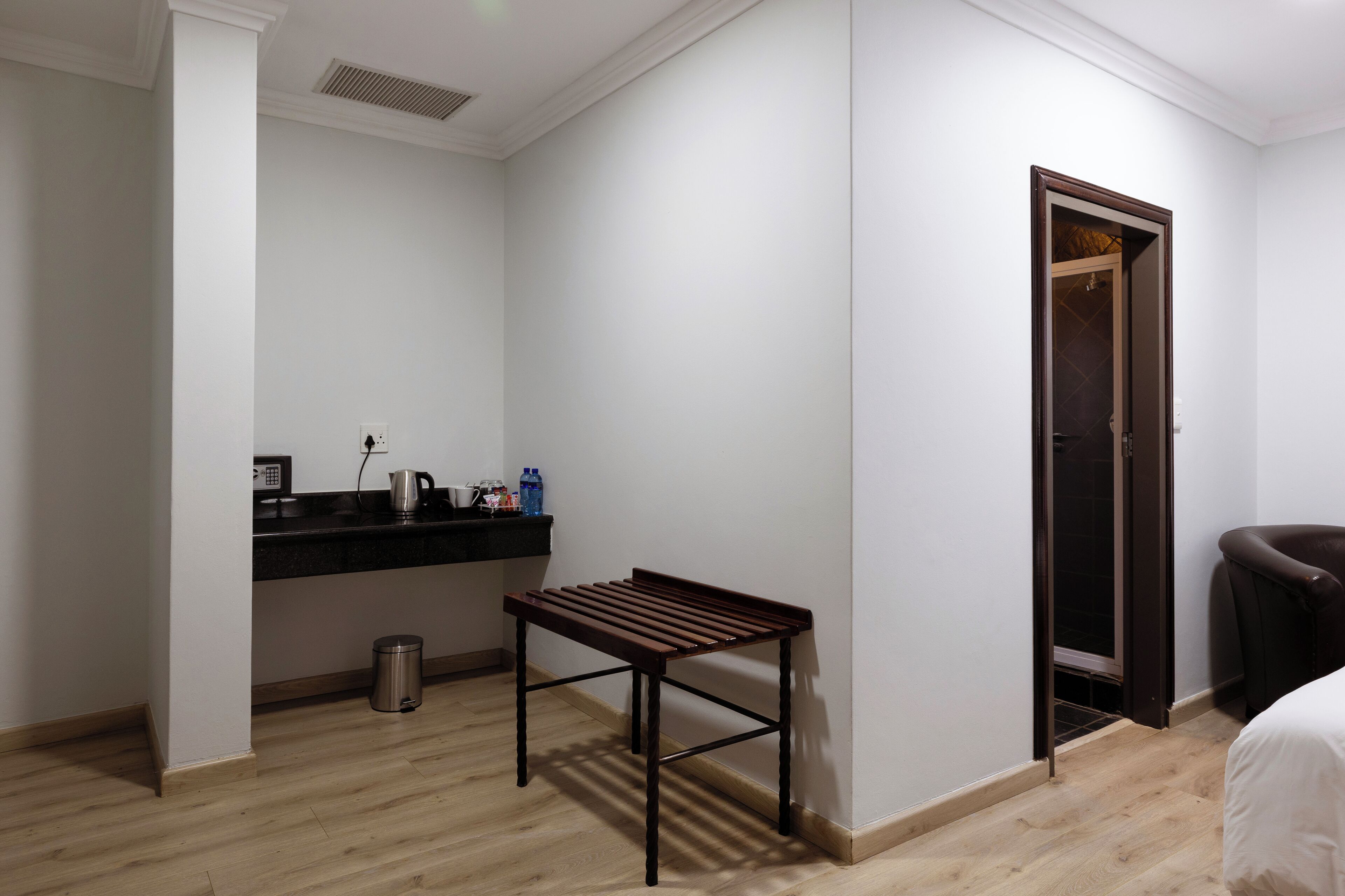 A hotel room with a wooden floor, a small table, a dark-colored chair, a coat rack, a door, and a black countertop with a kettle and bottles.