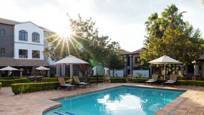 A swimming pool with lounge chairs and umbrellas, surrounded by buildings and trees. - Basdenwood Hotel  (Centurion)