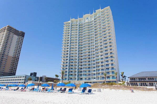 A high-rise hotel with multiple balconies, a beach area with sun loungers and umbrellas, and a clear blue sky.