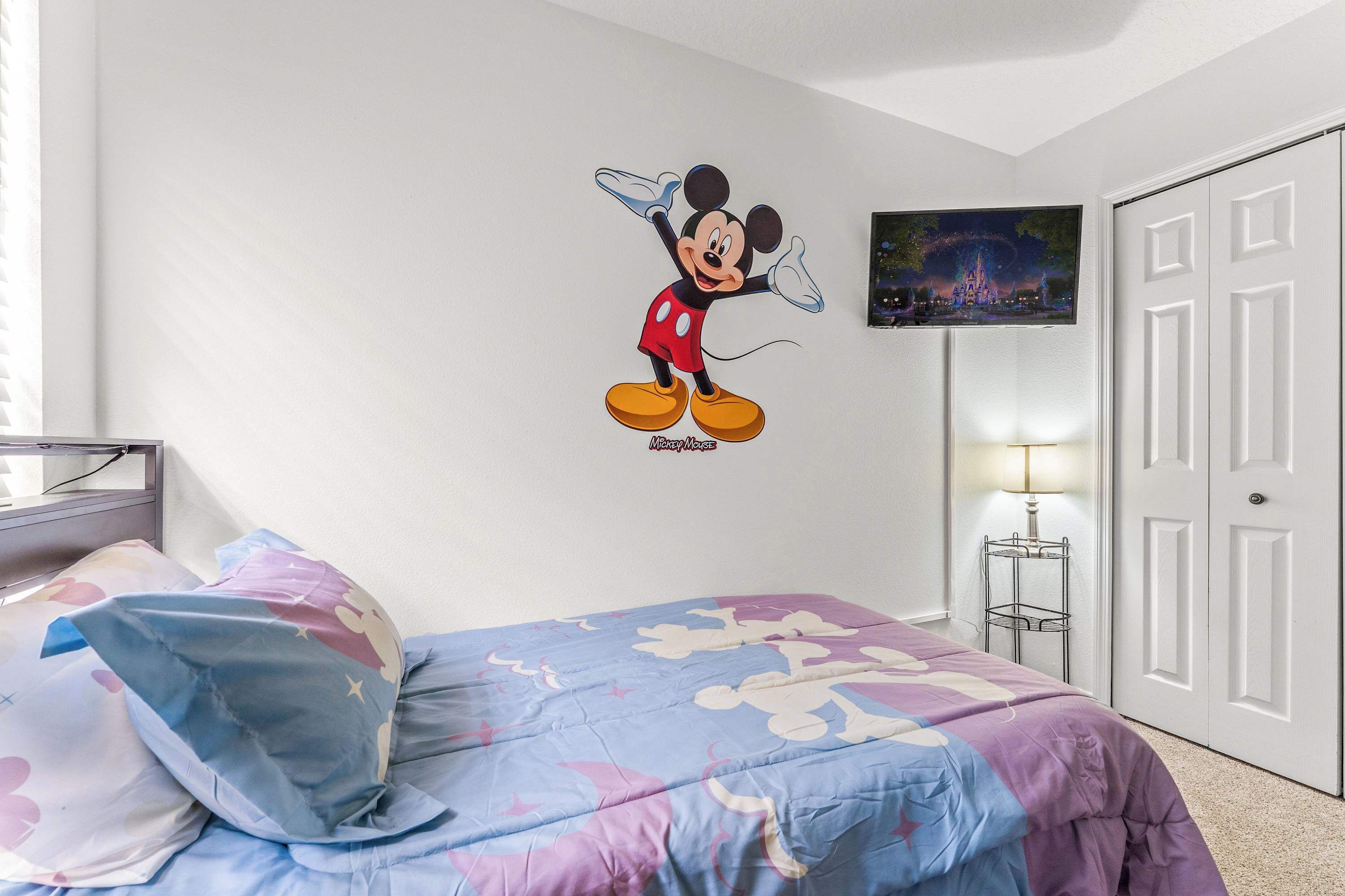 Et soveværelse med Mickey Mouse-vægdekoration, en seng med Mickey Mouse-dynebetræk, et natbord med lampe og en dør med et billede af et slot.