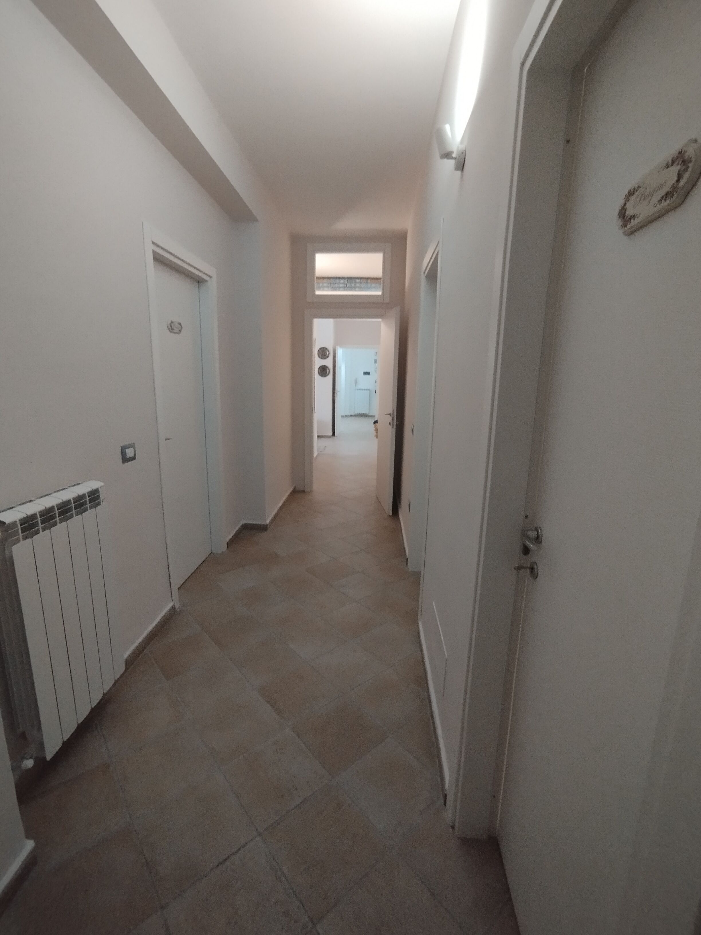A narrow hallway with white walls and tiled flooring, leading to a door at the end.