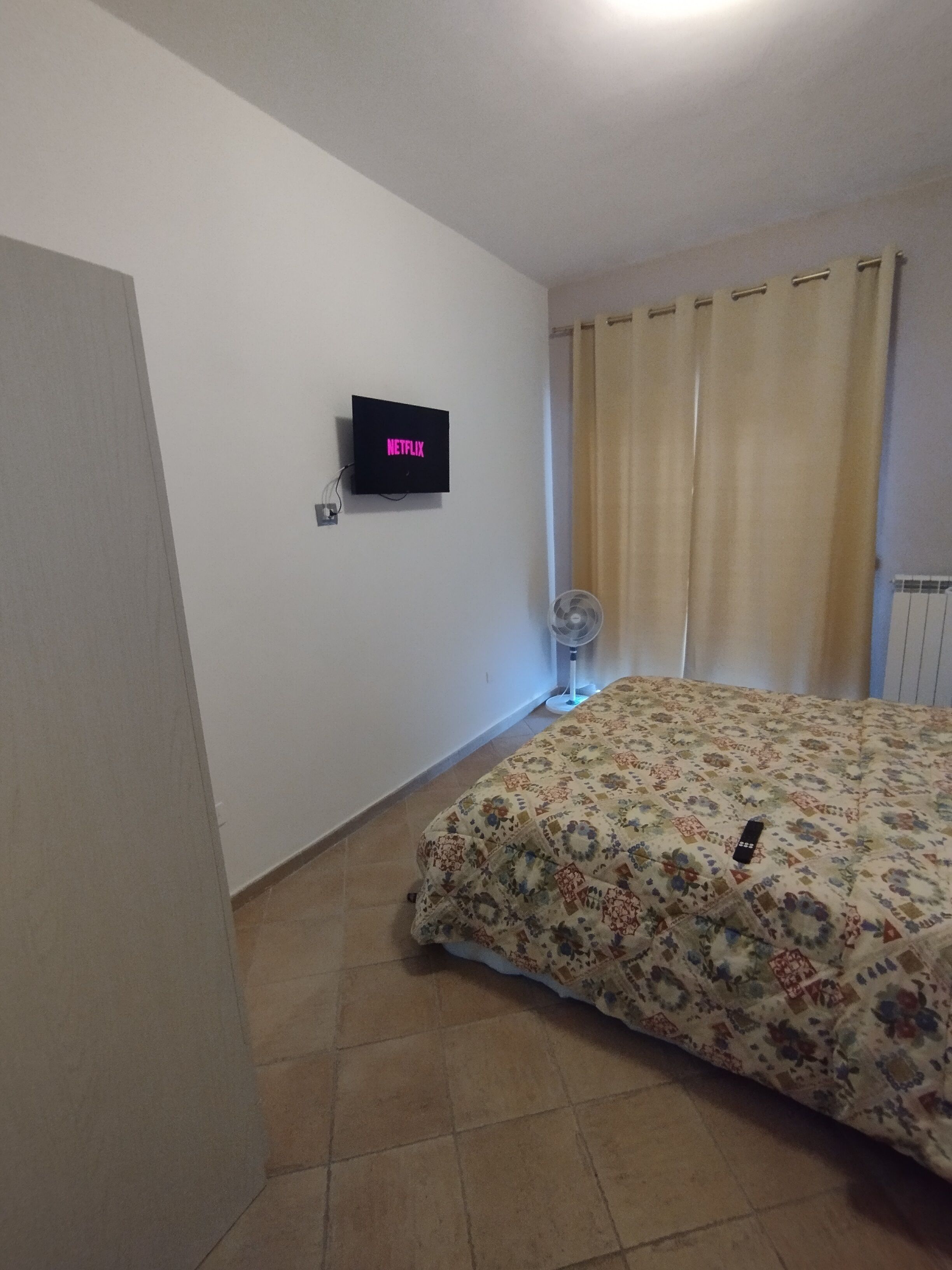 Quarto com cama, televisão na parede, ventilador e janela com cortinas.