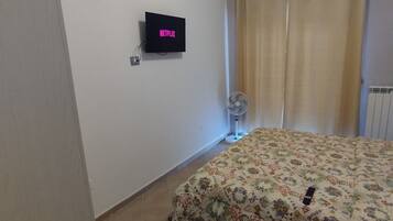 Quarto com cama, televisão na parede, ventilador e janela com cortinas.