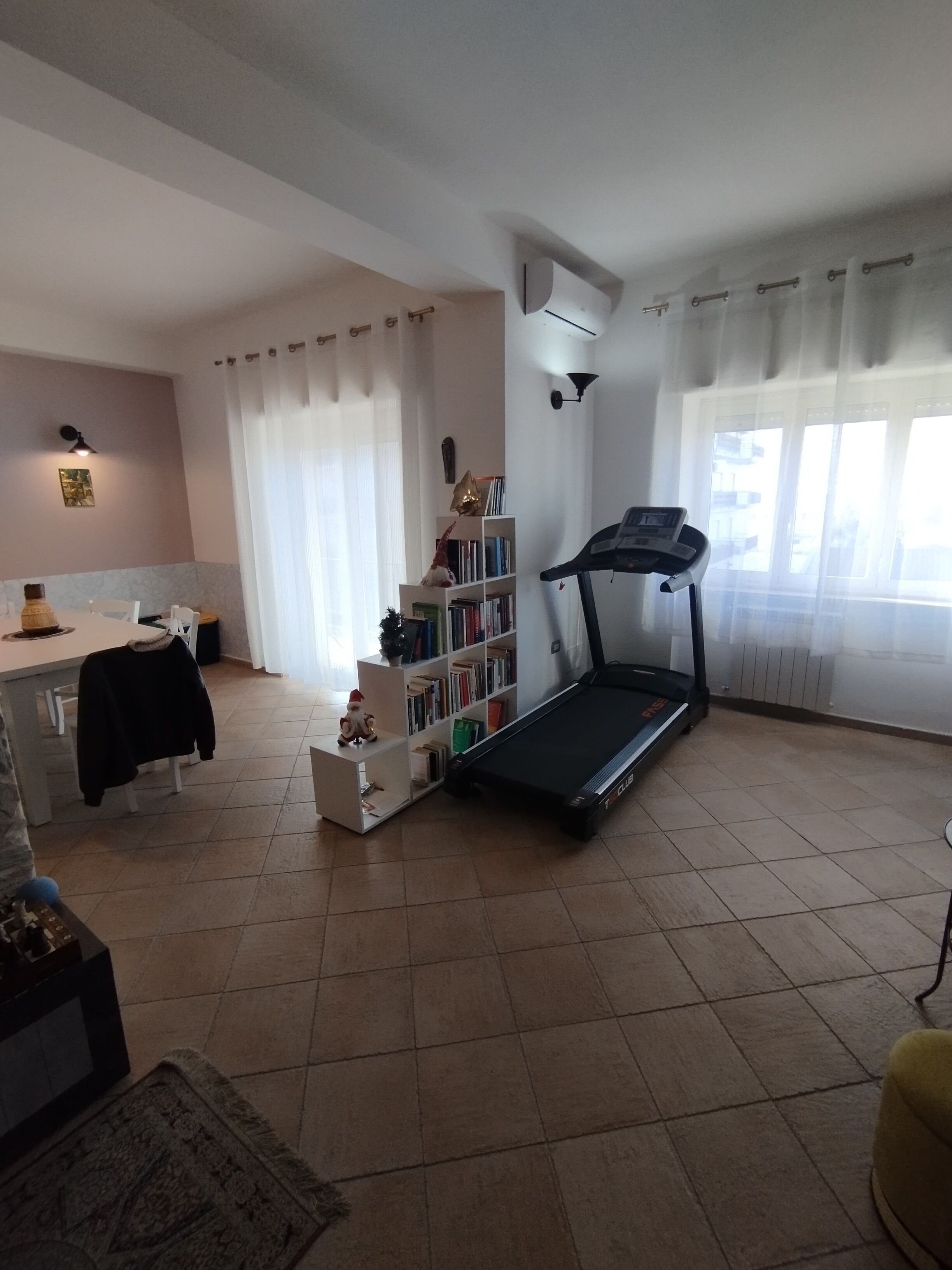 Una sala de estar con una caminadora, un comedor con mesa y sillas, una estantería y un ventanal con cortinas.