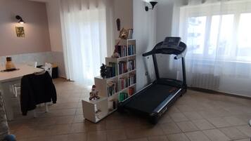 Una sala de estar con una caminadora, un comedor con mesa y sillas, una estantería y un ventanal con cortinas.