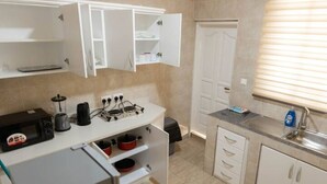 Une cuisine compacte avec des meubles blancs, un plan de travail muni d’une bouilloire, d’une machine à café et d’un four à micro-ondes. On y trouve une porte avec une fenêtre, ainsi qu’un évier avec un distributeur de savon.