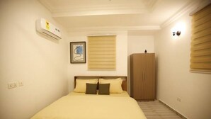 Une chambre à coucher comprenant un lit, un climatiseur, une fenêtre avec des stores, une armoire et un luminaire fixé au mur.