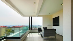Un balcón moderno con barandilla de vidrio, un sofá de mimbre y vistas a la ciudad.
