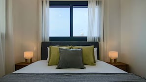 Una cama bien hecha con un cabecero oscuro, dos lámparas de noche y un ventanal con cortinas.